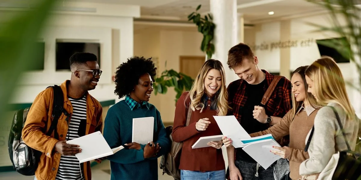 Zdjęcie ilustracyjne. Studenci i studentki stoją na korytarzu, w rękach trzymają notatniki.